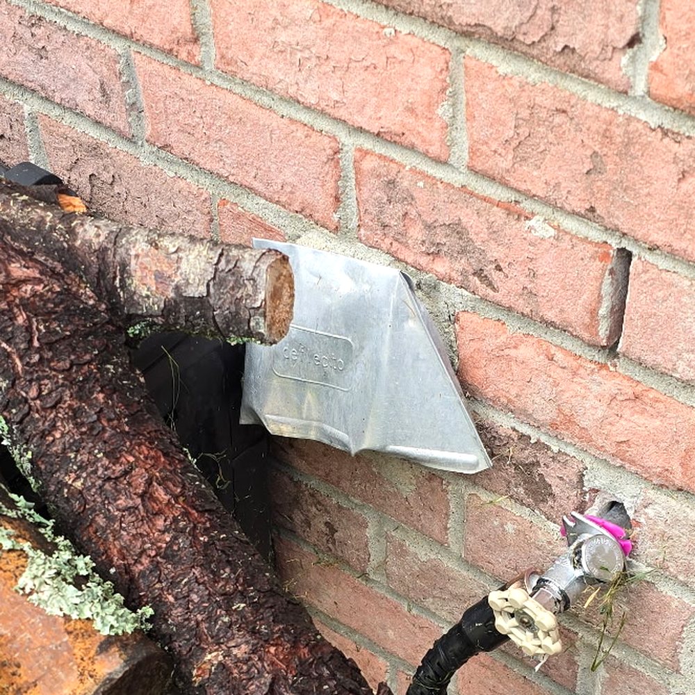 A crushed metal dryer vent hood on a brick wall, blocked by a stack of firewood